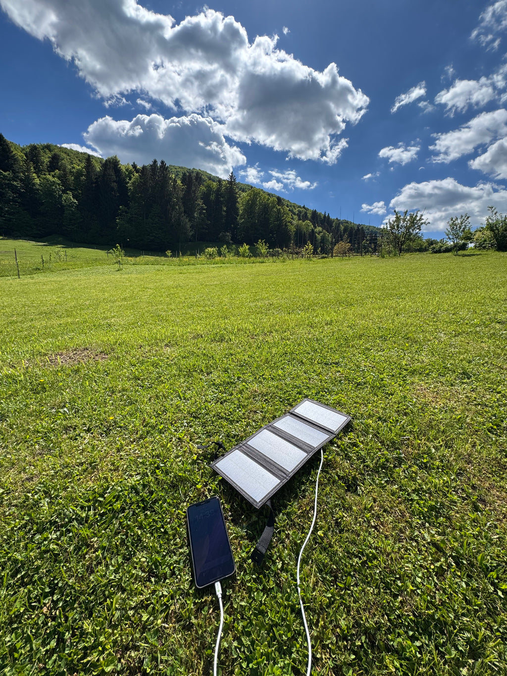 Solar panel charging a phone on a grassy field with trees and blue sky in the background